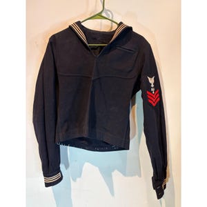 May include: A dark navy blue long-sleeve sailor jumper featuring a traditional square collar with white trim, matching white striped cuffs, and a military-style patch on the left sleeve displaying a white eagle emblem above three red chevrons.