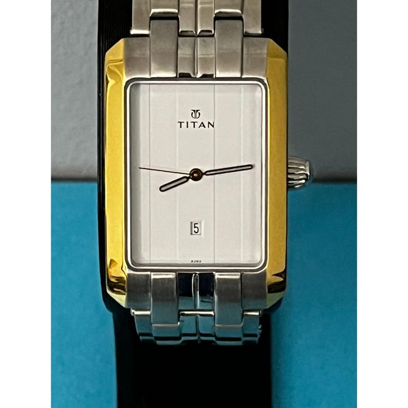 May include: A rectangular Titan wristwatch with a silver and gold-tone case and band. The watch face is white with black hands and the date displayed at the bottom. The watch is displayed on a black surface.