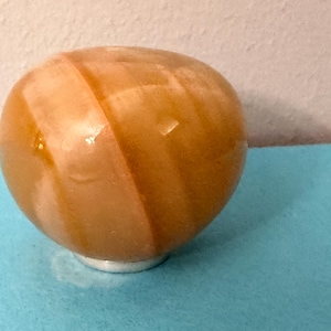 May include: A polished, spherical decorative object with a warm, amber color and a subtle, light-colored stripe. The sphere sits on a small, silver-colored base, resting on a blue surface. The object has a smooth, glossy finish.