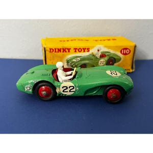 May include: A vintage green Dinky Toys race car model with the number 22 on the side and a white driver figure. The car has red wheels and is set against a yellow box with the Dinky Toys logo.