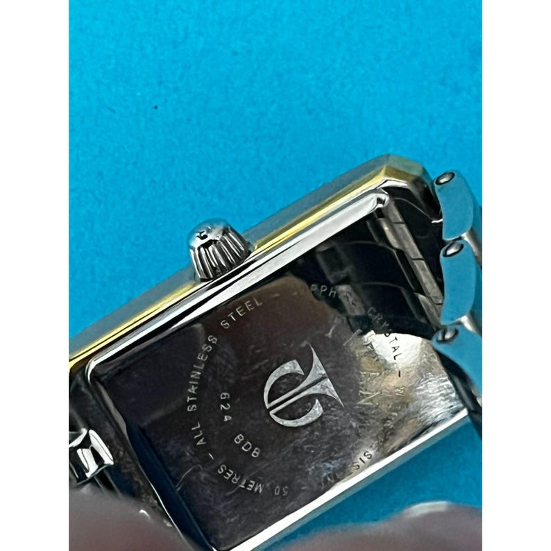 May include: A rectangular stainless steel watch with a silver band and gold accents. The watch face is not visible, but the back displays the brand logo, model number, and the words "ALL STAINLESS STEEL", "50 METRES", and "SAPPHIRE CRYSTAL".