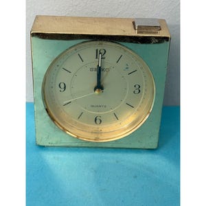 May include: A vintage Seiko quartz alarm clock with a gold-tone case and a light green face. The clock face has black numerals and hands, with the Seiko logo and "QUARTZ" text. The clock is rectangular with a small silver-tone button on top.