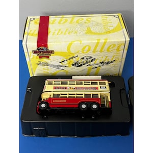 May include: A red and cream double-decker bus model with the words "Lion Black Lead" and "London United" on the side. The bus is in a black display case with a yellow and white box behind it.