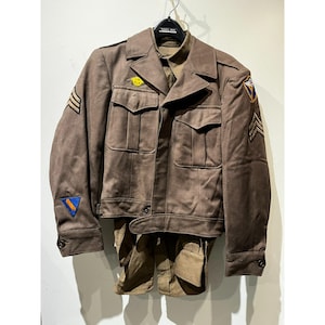May include: A vintage brown military uniform, including a jacket and trousers, hangs on a hanger. The jacket features multiple pockets, patches, and stripes. The uniform is a muted brown color, suggesting it is an older piece.