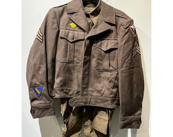 May include: A vintage brown military uniform, including a jacket and trousers, hangs on a hanger. The jacket features multiple pockets, patches, and stripes. The uniform is a muted brown color, suggesting it is an older piece.