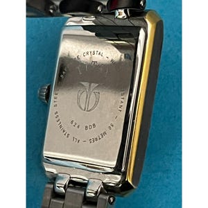 May include: Close-up of a rectangular wristwatch with a silver stainless steel band and case. The watch face is gold-toned. The back of the watch has the text "SAPPHIRE CRYSTAL - WATER RESISTANT - ALL STAINLESS STEEL - 50 METRES".