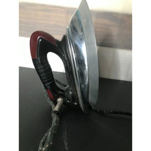 May include: A vintage black and silver iron with a red handle and a cord. The iron has a metal soleplate and a cord that is wrapped around the handle.