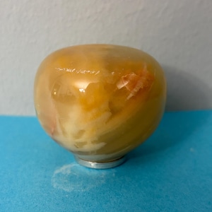 May include: A decorative, rounded object with a translucent, amber-colored exterior. The interior has a mix of yellow and white hues, with a small, silver-colored base. The object is set against a blue surface.