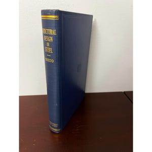 May include: A hardcover book titled "Structural Design in Steel" by Shedd. The book has a dark blue cover with gold lettering and a gold-colored top edge. The book is standing upright on a wooden surface.