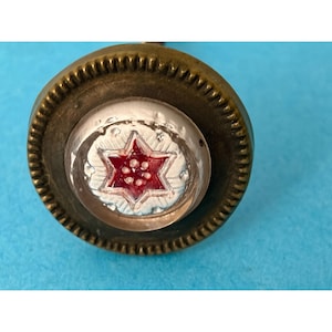 May include: A round brass button featuring a textured, ridged outer edge and a central circular inlay. The inlay displays a clear, domed surface over a white background with a six-pointed star design in red, accented by small white dots.