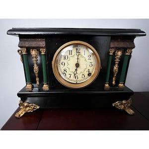 May include: A black antique mantel clock with a gold-rimmed clock face. The clock has gold accents, green columns, and ornate gold feet. The clock face displays black numerals and hands.