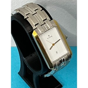 May include: A rectangular Titan wristwatch with a silver band and gold-toned accents. The watch face is white with black hands and the date displayed at the bottom. The watch is displayed on a black stand.