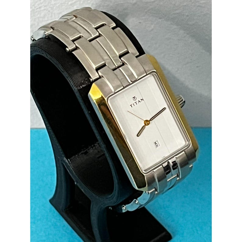 May include: A rectangular Titan wristwatch with a silver band and gold-toned accents. The watch face is white with black hands and the date displayed at the bottom. The watch is displayed on a black stand.