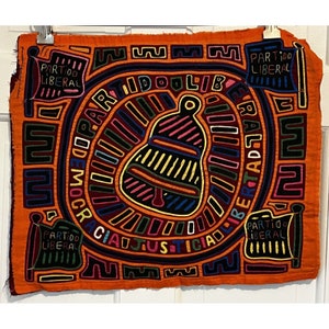 May include: A colorful mola textile with a bell design in the center. The textile is orange with black, yellow, blue, green, and red details. The words "Partido Liberal" are repeated around the border.