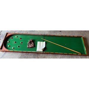 May include: A vintage tabletop game with a green felt playing surface, wooden frame, and a curved end. Includes a cue stick, small balls, a drawstring bag, and a paper sheet. The game is set on a concrete surface.