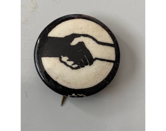 May include: Black and white button with a graphic of two hands shaking. The button is round and has a black border.
