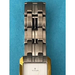 May include: A rectangular wristwatch with a silver-tone metal band and a gold-tone bezel. The watch face is white with the brand name "TITAN" printed in black. The watch band is made of interlocking rectangular links.