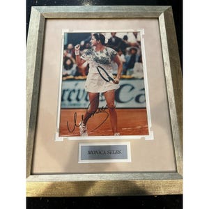 May include: Framed photograph of Monica Seles, a tennis player, in action on a clay court. She is wearing a white tennis outfit and holding a racket. The photo is signed and includes a nameplate that reads "Monica Seles."