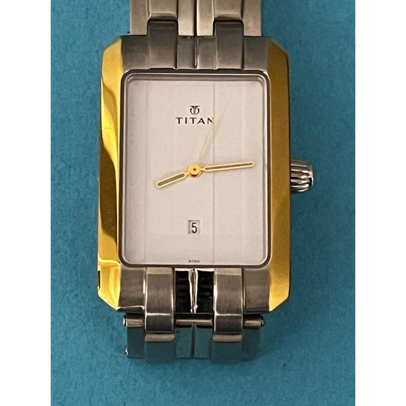May include: A rectangular Titan wristwatch with a silver and gold-tone metal band. The watch face is white with gold hands and a date display at the 6 o'clock position. The watch has a gold-tone bezel and a silver-tone metal band.
