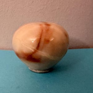 May include: A small, rounded, light-colored decorative object with brown marbling. The object has a smooth, glossy surface and sits on a small, circular base. The background is a light pink wall and a blue surface.