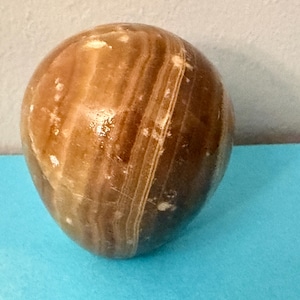 May include: A polished, egg-shaped stone with a warm, brown color and a striped pattern. The stone is set against a blue surface, creating a contrast that highlights its natural beauty. This decorative item could be used as a paperweight or a unique home decor piece.