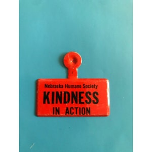 May include: Red metal tag with a black inscription that reads "Nebraska Humane Society Kindness in Action".