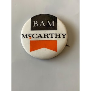 May include: A white button with a black and orange design. The button says "BAM MCCARTHY" with a star between the words.