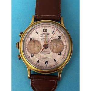 May include: A vintage gold-tone Aureolf Chronograph watch with a cream-colored face, featuring two sub-dials and a brown leather strap. The watch face displays Arabic numerals and the text "AUREOLF CHRONOGRAPHE". The watch has a classic design.