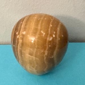 May include: A polished, egg-shaped stone with a warm, amber hue. The surface features natural, light brown and white veining, creating a striped pattern. The stone is smooth and rounded, with a slight sheen.