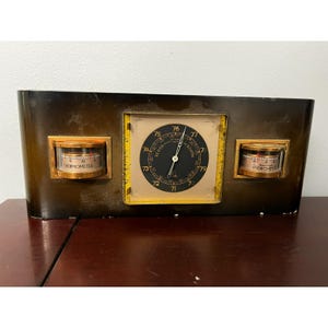 May include: A vintage weather station with a dark brown, curved frame. It features a central clock, a thermometer on the left, and a hygrometer on the right. The clock face is black with yellow accents, and the thermometer and hygrometer have gold-colored frames.