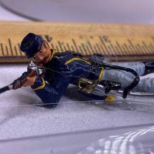 Puede incluir: Una figurita de soldado de hojalata pintada con un uniforme azul con ribetes amarillos, sosteniendo un rifle y acostado en posición de decúbito prono.