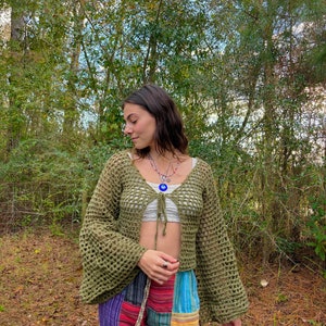 K&ouml;nnte beinhalten: Eine gr&uuml;ne H&auml;keljacke mit Bindeband vorne und Glocken&auml;rmeln. Die Jacke wird &uuml;ber einem wei&szlig;en Crop-Top und einer bunten Patchworkhose getragen.