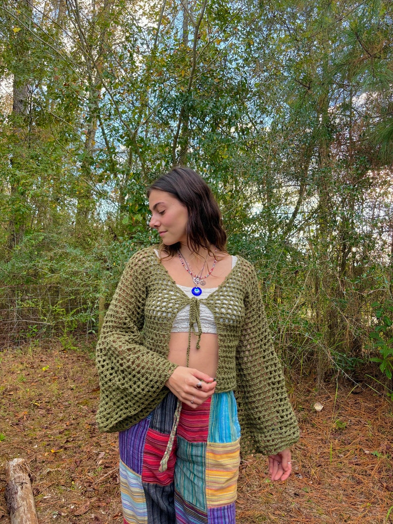 K&ouml;nnte beinhalten: Eine gr&uuml;ne H&auml;keljacke mit Bindeband vorne und Glocken&auml;rmeln. Die Jacke wird &uuml;ber einem wei&szlig;en Crop-Top und einer bunten Patchworkhose getragen.