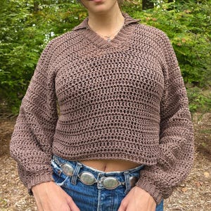 May include: A brown, open-weave crochet sweater with a V-neck collar and long sleeves. The cropped sweater is paired with blue denim jeans and a silver belt. The sweater's texture and color suggest a handmade or artisanal quality.