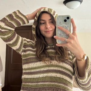 Pode incluir: Um moletom com capuz de crochê em um padrão listrado de verde oliva, marrom e creme. O moletom com capuz tem um capuz com cordão e mangas compridas com punhos flare. A peça de roupa é feita de um fio macio e texturizado.