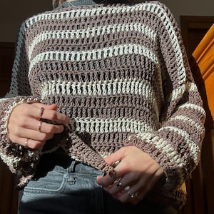 Puede incluir: Un su&eacute;ter de crochet a rayas marrones y blancas con un corte holgado y mangas largas.