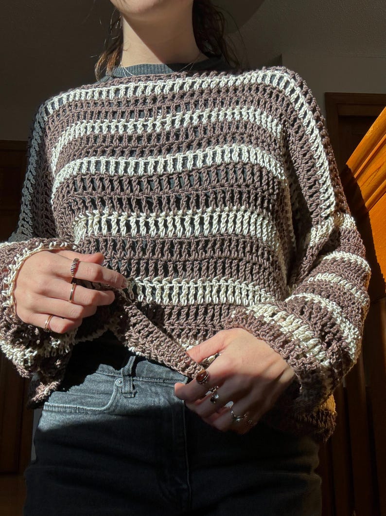 Puede incluir: Un su&eacute;ter de crochet a rayas marrones y blancas con un corte holgado y mangas largas.