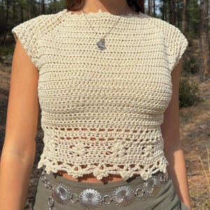 Puede incluir: Top de crochet color crema, hecho a mano, con un patrón floral en la cintura. El top tiene mangas cortas y cuello redondo. Un cinturón de cadena plateada con medallones decorativos y pantalones verde oliva completan el look.