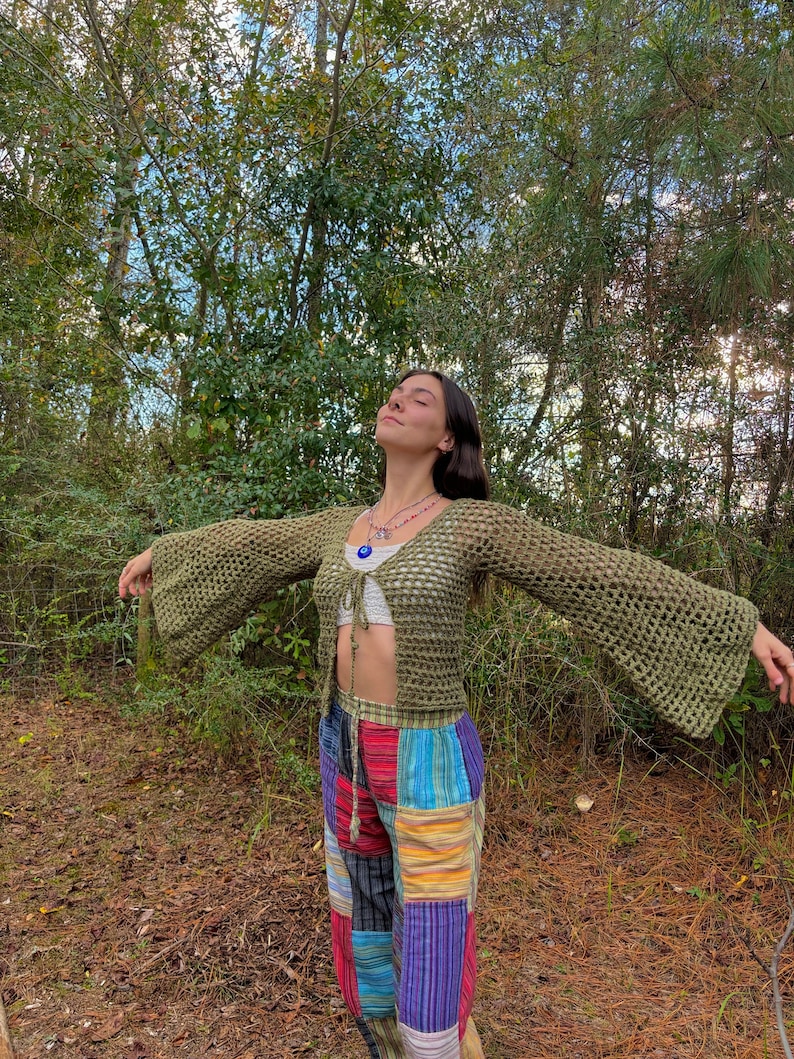 K&ouml;nnte beinhalten: Eine Frau tr&auml;gt ein gr&uuml;nes H&auml;keltop mit Glocken&auml;rmeln und eine bunte Patchworkhose. Sie steht in einem Wald mit gr&uuml;nen B&auml;umen und braunen Bl&auml;ttern auf dem Boden.