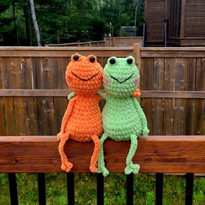 May include: Two crocheted frog plushies sitting on a wooden railing. The frog on the left is orange and the frog on the right is green.
