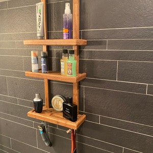 Puede incluir: Una estantería de madera con tres estantes, montada en una pared de azulejos. Los estantes están llenos de diversos artículos de tocador, incluyendo botellas de gel de ducha, laca para el cabello y colonia. También hay dos maquinillas de afeitar colgadas del estante inferior.