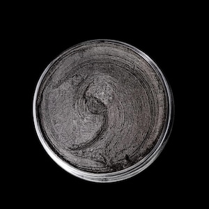Puede incluir: Un tarro redondo transparente lleno de un producto cosmético gris oscuro. La superficie del producto tiene una textura y un patrón arremolinado. El fondo es negro. El producto parece ser una crema o pasta.