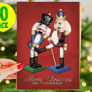 May include: A Christmas card featuring two nutcracker figurines. The nutcracker on the left is wearing a blue uniform and is being kicked in the rear by the nutcracker on the right, who is wearing a red uniform. The card has a red background and the text "Merry Christmas THE NUTCRACKER".