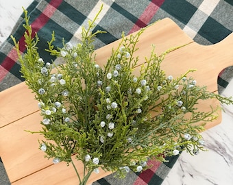 May include: A sprig of green faux juniper with small white berries rests on a wooden cutting board. The board is placed on a green, red, and white plaid patterned cloth. The background is a white marble surface.
