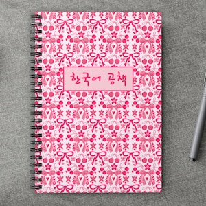 May include: A spiral-bound notebook with a pink and white floral pattern featuring bows, cherries, and flowers. The notebook has the text "한국어 공책" in a pink rectangle.