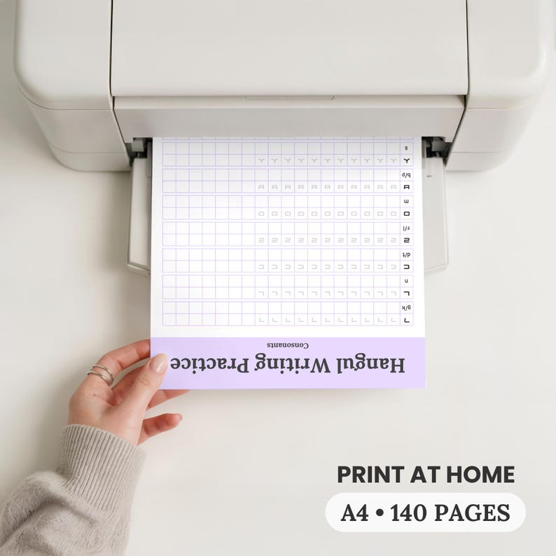 A person in a cozy sweater pulls an A4 Hangul writing practice page from a home printer, highlighting that the 140 page Korean workbook is easy to print at home for handwriting practice. Keywords: printable Korean workbook, Hangul practice sheets, A4 PDF, home printing.