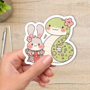 A sticker with a green snake with pink flowers and a gray rabbit wearing a pink kimono. The snake has a green and pink floral pattern and the rabbit has pink cheeks.
