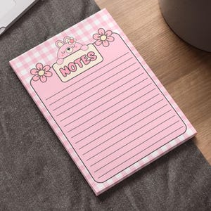 May include: Pink notepad with a gingham border and lined pages. A cartoon bear holds a sign that says "NOTES". The notepad has a pink and white color scheme with flower accents.