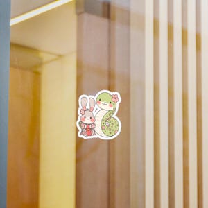A sticker of a green snake with pink flowers and a white rabbit wearing a red and white kimono. The snake and rabbit are facing each other and the snake is wrapped around the rabbit.