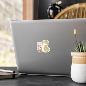 A sticker with a cartoon illustration of a green snake with pink flowers and a white rabbit wearing a pink dress and a pink flower in its hair.